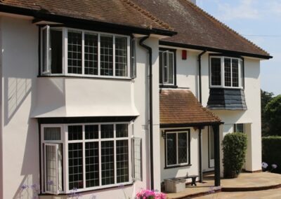 Two‑storey white house with large bay heritage windows, black trim and potted plants along the paved frontage.