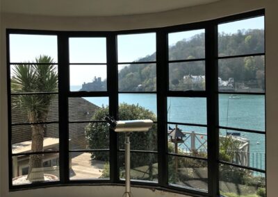 Large curved black‑framed window overlooking a waterfront landscape with hills and historic buildings.